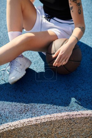 Una mujer joven se sienta en el suelo con una pelota de baloncesto, perdida en el pensamiento y lista para jugar.