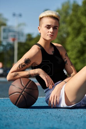 A young woman with tattoos sits on the ground, holding a basketball, lost in thought.