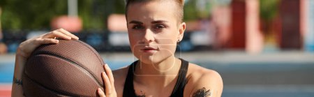Photo for A young woman with short hair and tattoos confidently holds a basketball in her right hand, ready to showcase her athletic skills outdoors. - Royalty Free Image