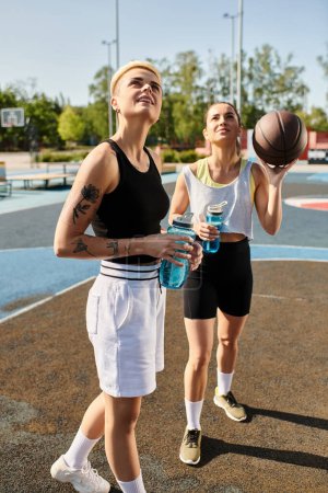 Dos mujeres jóvenes de pie con confianza en una cancha de baloncesto, exudando fuerza y determinación en atuendo deportivo en un día soleado.