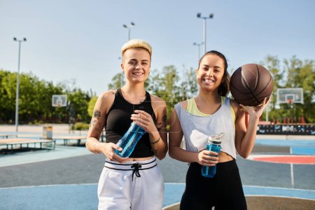Two young women, holding water bottles, prepare to play basketball outdoors on a sunny day in summer.