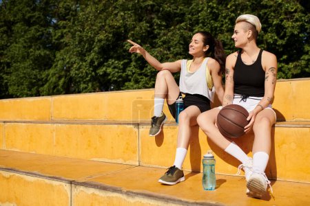 Two young women, athletic friends, sit closely together after playing basketball outdoors on a summer day.