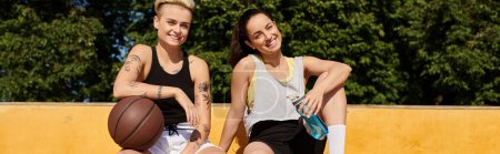 Zwei junge Frauen in Sportkleidung sitzen auf einem Basketballfeld, unterhalten sich und genießen eine entspannende Pause nach einem Spiel.
