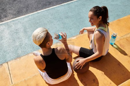Zwei athletische junge Frauen sitzen auf dem Boden und trinken mit Wasserflaschen, nachdem sie im Sommer draußen Basketball gespielt haben.