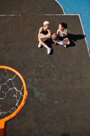 Two young women sit on top of a basketball court, chatting and enjoying the summer day.