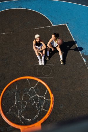 A pair of athletic female friends sitting on top of an outdoor basketball court on a sunny day.