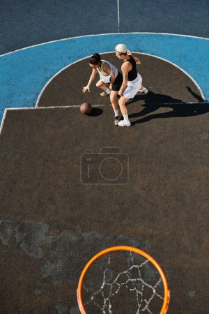 Zwei junge Frauen in Sportbekleidung spielen leidenschaftlich Basketball auf einem sonnigen Außenplatz.