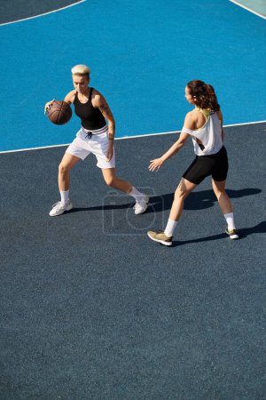Two young women, athletic and competitive, playing basketball on an outdoor court on a summer day.