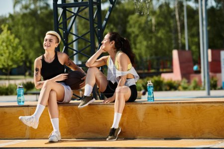 Zwei athletische junge Frauen sitzen auf einem Felsvorsprung und teilen einen Moment der Freude und des Lachens, während sie einen Sommertag im Freien genießen.