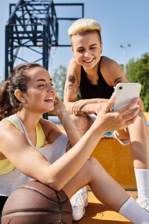 Dos mujeres jóvenes atléticas, sentadas en el suelo al aire libre, enfocadas en la pantalla de un teléfono inteligente.
