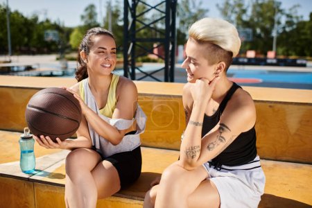 Téléchargez les photos : Deux femmes athlétiques assises côte à côte, tenant un ballon de basket, profitant d'une journée d'été en plein air. - en image libre de droit