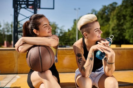 Zwei junge Frauen, Freundinnen, sitzen auf einem Basketballfeld, die Arme umeinander, genießen ihre gemeinsame Zeit an einem sonnigen Tag.