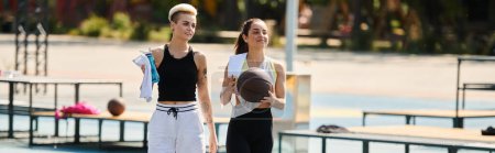 Dwie młode kobiety grają w koszykówkę na boisku, pokazując swoje umiejętności sportowe i pracę zespołową w letnim słońcu.