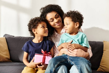 Téléchargez les photos : Gaie femme afro-américaine étreignant ses adorables petits fils qui tiennent présent, Fête des mères - en image libre de droit