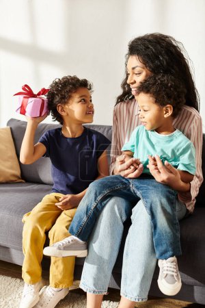 fröhliche afrikanisch-amerikanische Frau, die ihren Sohn hält und ein Geschenk von einem anderen Sohn erhält, Muttertag