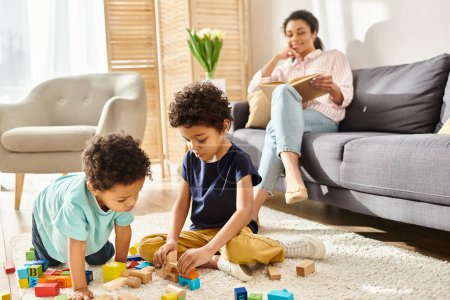 Foto de Se centran en chicos afroamericanos lindos jugando juguetes con su borrosa madre leyendo el libro sobre el telón de fondo - Imagen libre de derechos