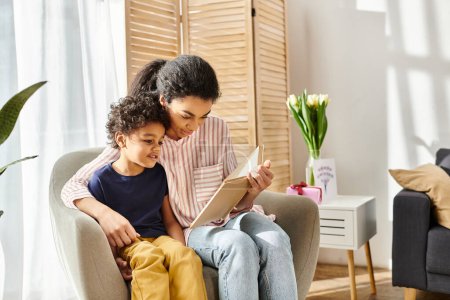 atractiva madre afro-americana alegre en traje acogedor leyendo interesante libro a su pequeño hijo