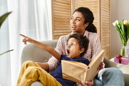 surprised little african american boy pointing away while reading book with his cheerful mother