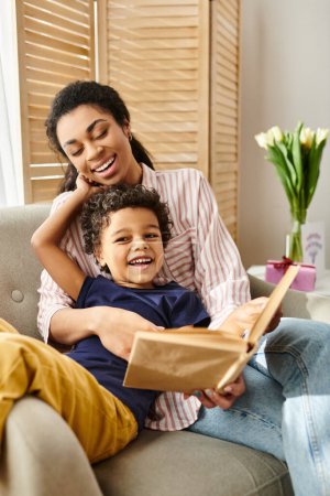 alegre afroamericana bonita mujer en la ropa de casa y su adorable pequeño hijo leer libro juntos