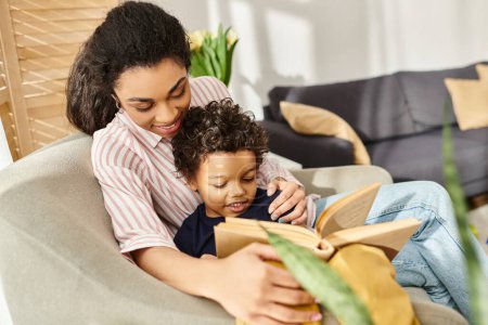 alegre cariñosa mujer afroamericana en ropa de casa abrazando a su pequeño hijo y leyendo libro con él