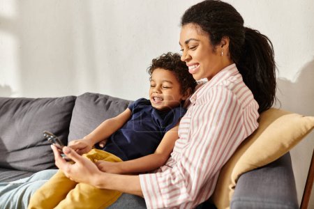 alegre hermosa africana americana madre y su pequeño hijo mirando el teléfono mientras está sentado en sofá
