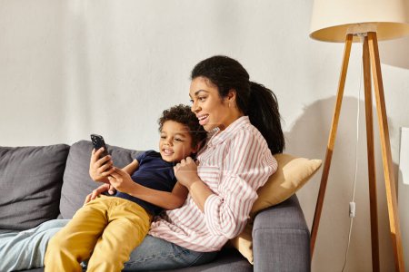atractiva madre afroamericana alegre y su adorable hijo sentado en el sofá y mirando el teléfono