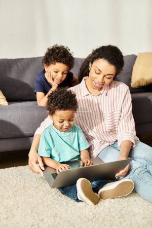 Téléchargez les photos : Joyeuse mère afro-américaine en tenue de maison profitant des films sur ordinateur portable avec ses deux fils mignons - en image libre de droit