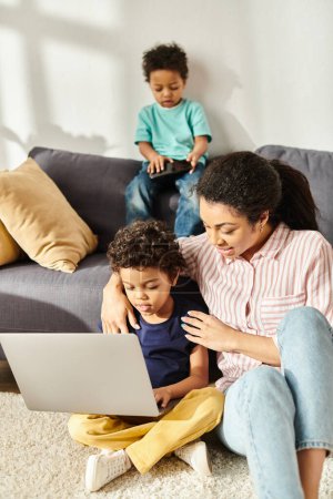 Téléchargez les photos : Joyeuse femme afro-américaine passe du temps avec ses deux fils joyeux et mignons tenant ordinateur portable et téléphone - en image libre de droit