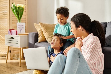 joyous african american mother in homewear looking with love at her little cute sons with laptop