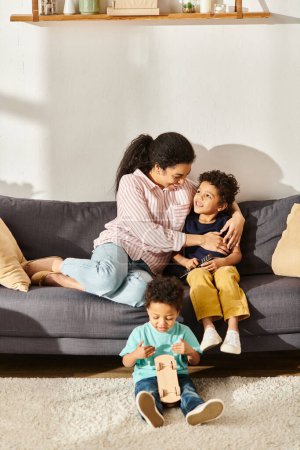 Téléchargez les photos : Joyeuse mère afro-américaine assise à regarder des films avec ses adorables fils dans le salon à la maison - en image libre de droit