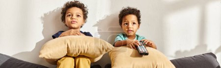 Téléchargez les photos : Adorables garçons afro-américains assis sur un canapé avec des oreillers et regarder des films intéressants - en image libre de droit