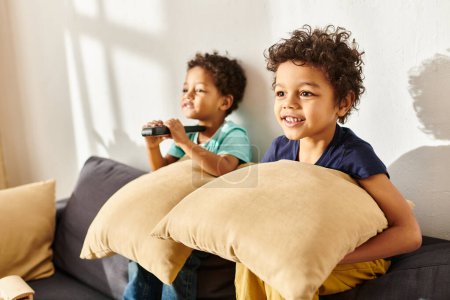 foco en alegre afroamericano chico con almohada viendo la televisión al lado de su borrosa adorable hermano