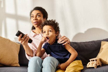 madre e hijo afroamericanos sorprendidos sentados en el sofá y viendo películas, crianza moderna