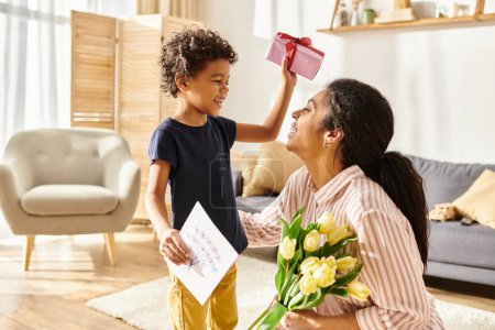 adorable afroamericano chico dando regalo y postal a su alegre madre, crianza moderna