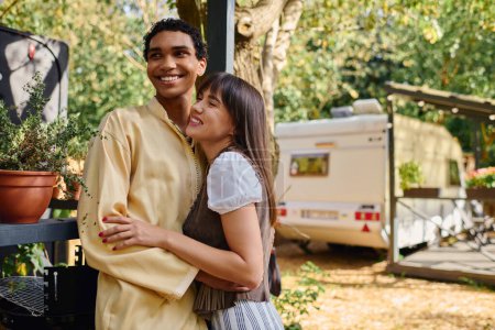 Foto de Un hombre y una mujer se abrazan calurosamente frente a una caravana, disfrutando de una escapada romántica en un entorno natural. - Imagen libre de derechos