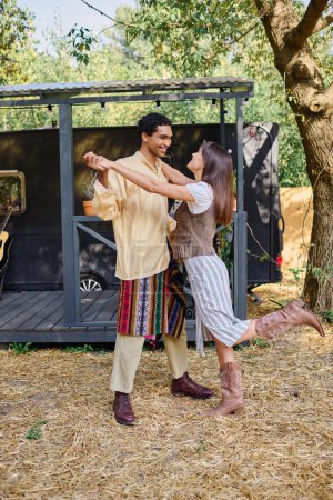 A man and woman of different races sway gracefully, dancing in front of their charming camper trailer in a scenic natural setting.