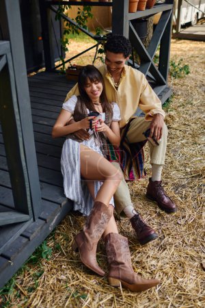 Téléchargez les photos : Un couple interracial assis paisiblement ensemble sur un porche en bois, entouré par la nature. - en image libre de droit