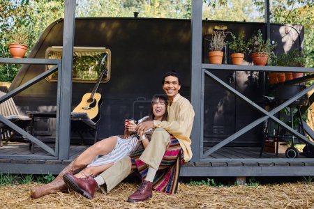 An interracial couple sits on the ground in front of a trailer, enjoying a romantic getaway in a serene natural environment.