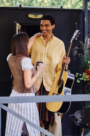 A man serenades a woman with a guitar in a natural setting near a camper van, creating a romantic atmosphere.