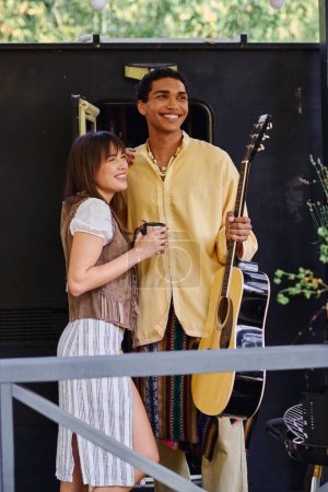 A man holds a guitar while standing next to a woman in a scenic outdoor setting, creating a harmonious moment of music and love.