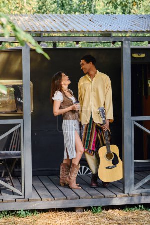 Una pareja interracial, hombre y mujer, de pie junto con una guitarra en la mano, ambos rodeados de naturaleza.