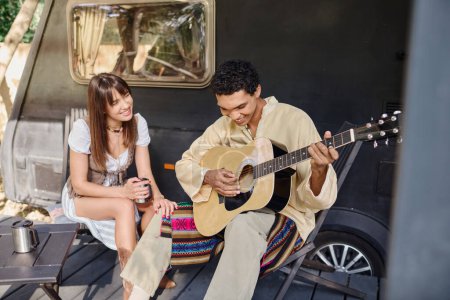 A man playing a guitar next to a woman in a romantic setting surrounded by nature, enjoying each others company.