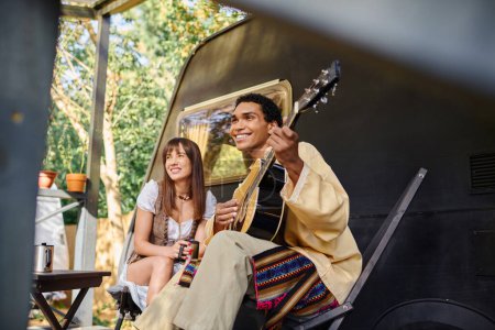 A man strums a guitar while sitting next to a woman, creating a harmonious melody by the campfire in a natural environment.