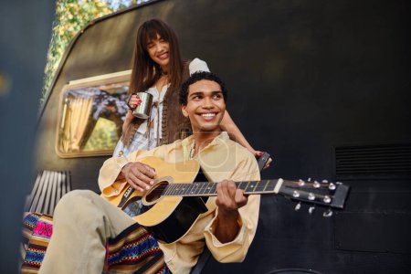A man serenades a woman with acoustic guitar music by a crackling bonfire in a serene outdoor setting.