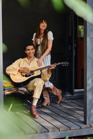 A man and a woman sit on a porch, strumming a guitar and enjoying each others company in a serene setting.