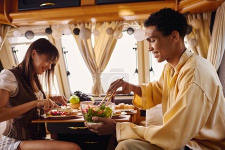 An interracial couple enjoying a delicious meal together inside a camper van during a romantic getaway.