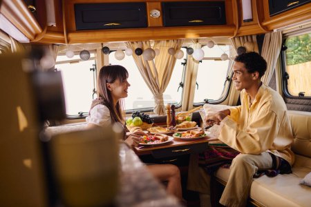 A man and a woman of different races enjoy a romantic lunch together inside a camper van during their travels.