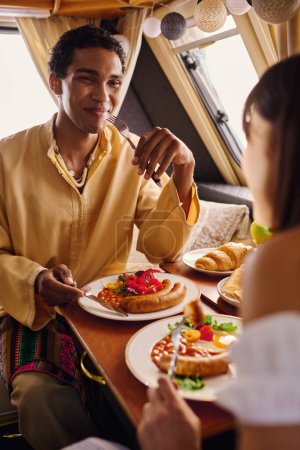 An interracial couple enjoying a romantic lunch together in a cozy camper van with plates of delicious food on the table.