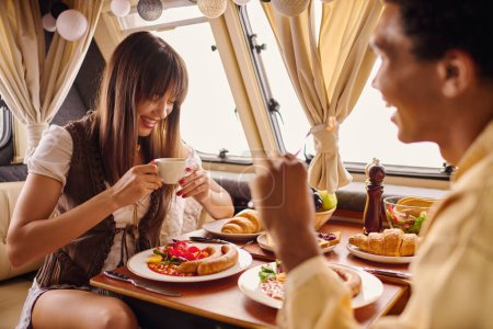 A man and a woman sit at a table with plates of food, enjoying a romantic lunch in a cozy camper van.
