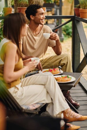Téléchargez les photos : Un couple interracial s'assoit sur un porche, mangeant de la nourriture ensemble dans un moment paisible et romantique pendant leur escapade. - en image libre de droit